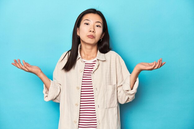 Asiatische Frau in geschichtetem Hemd und gestreiftem T-Shirt zweifelt und zuckt mit den Schultern