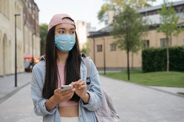 Asiatische Frau in medizinischer Maske, die Handy hält und im Freien wegschaut