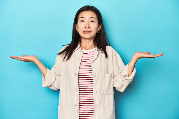 Asiatische Frau in mehrlagigem Hemd und gestreiftem T-Shirt zuckt verwirrt und zweifelnd mit den Schultern