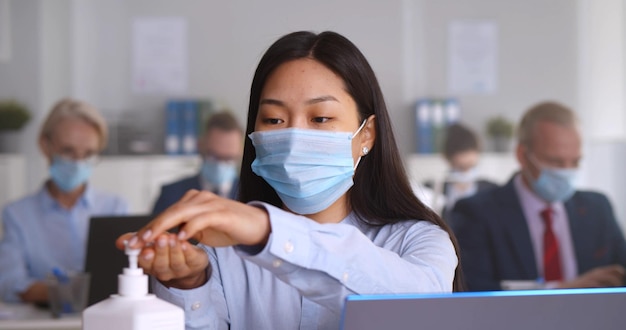 Asiatische Frau in Sicherheitsmaske, die im Büro die Hände mit antibakteriellem Gel reinigt