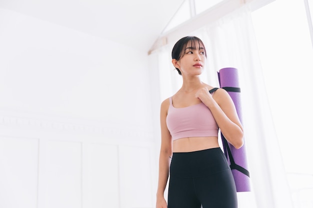 Asiatische Frau in Sportbekleidung und Yogahosen mit einer aufgerollten Yogamatte, die in einem Schlafzimmer mit Sonnenlicht aus dem Fenster steht Bild in voller Länge mit Kopierbereich