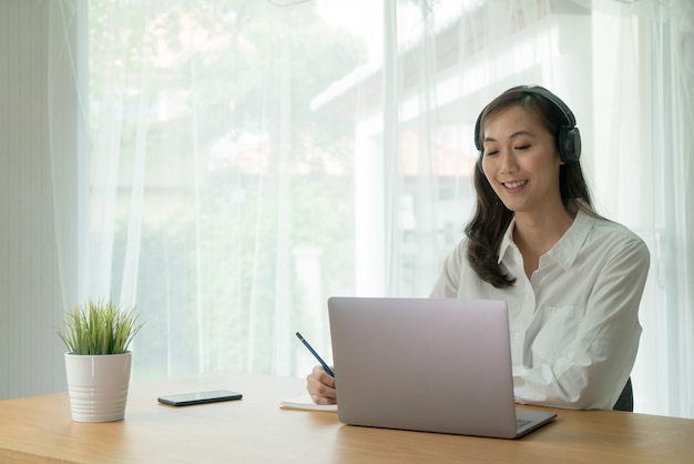 Asiatische Frau Lächeln und Videoanruf online vom Laptop-Notebook-Computer, der kabellosen Kopfhörer warnt und Notizen am Schreibtisch macht.