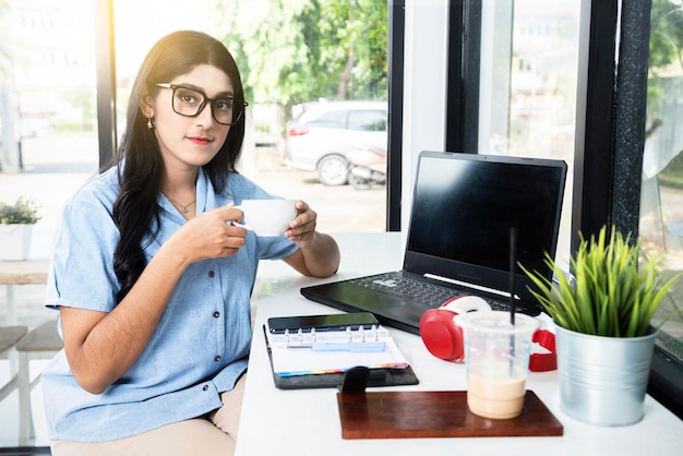 Asiatische Frau mit Brille mit einem Laptop und einem Notizbuch auf dem Tisch mit einer Tasse Kaffee