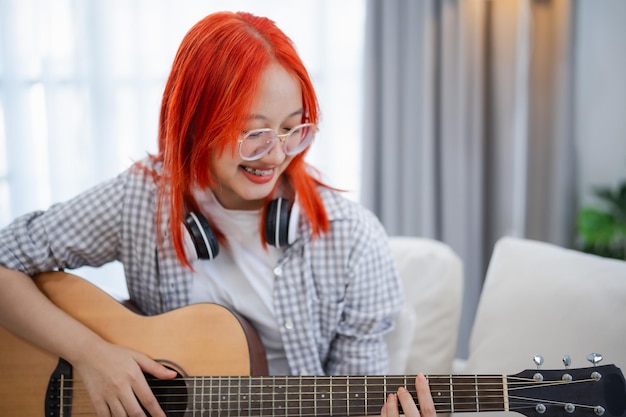 Asiatische Frau mit Brille und Kopfhörer spielt Gitarre, während sie im Wohnzimmer zu Hause auf dem Sofa sitzt Asiatische Frauen schreiben Lieder, während sie zu Hause Gitarre spielen Komponieren Sie Lieder Musikkonzept