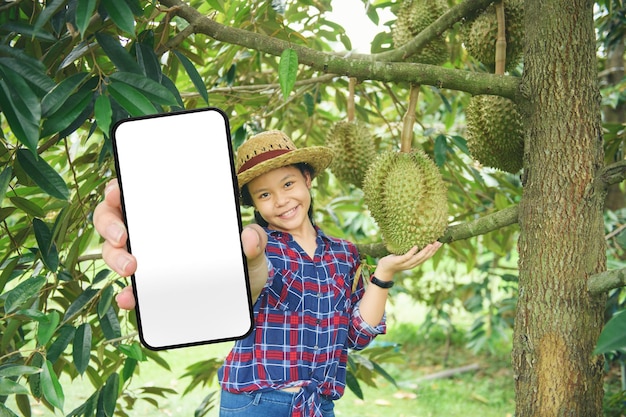 Asiatische Frau mit Hut in rot gestreiftem Hemd posiert mit Smartphone, das einen leeren Bildschirm im Durian Farm Garden zeigt Asia Girl Show Handy Display Mock Up