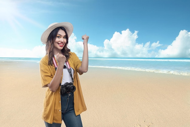 Asiatische Frau mit Hut und Kamera mit einem aufgeregten Ausdruck, der am Strand unterwegs ist