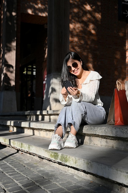 Asiatische Frau mit ihren Einkaufstaschen mit ihrem Smartphone, während sie an der Straßentreppe sitzt