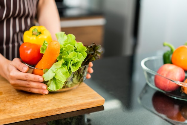 Asiatische Frau mit Schürze, die einen gesunden Salat mit Gemüse in einer Glasschüssel zubereitet.