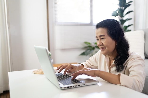 Asiatische Frau mittleren Alters Arbeit von zu Hause aus lächelnde ältere Frau Schöne reife asiatische Frau auf Laptop mit Technologiekonzept