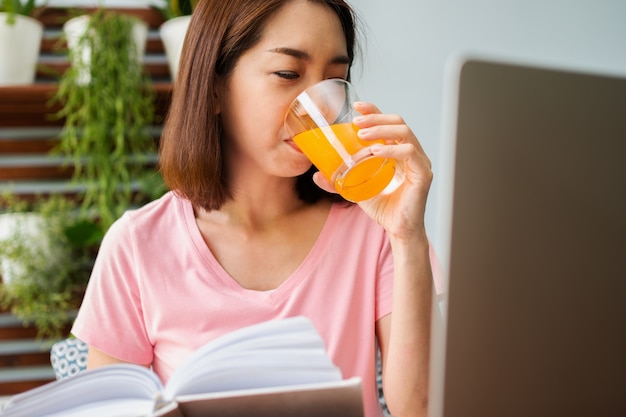 Asiatische Frau mittleren Alters, die ein Buch liest und Orangensaftglas im Haus hält. Konzept der Gesundheitsversorgung und gesunde Ernährung