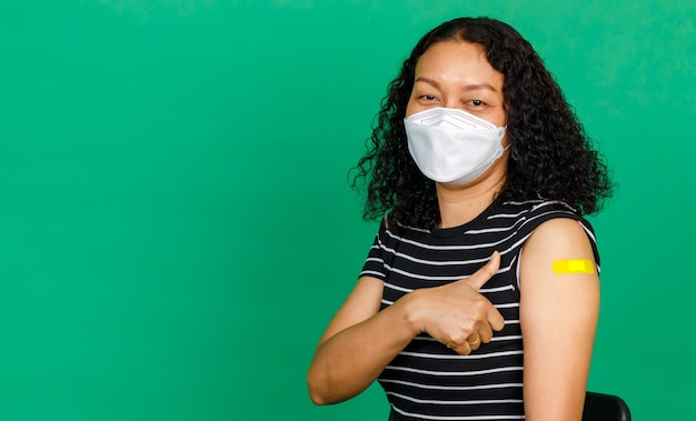 Asiatische Frau mittleren Alters mit Maske und Daumen hoch, ihr Arm mit Verbandspflaster zeigt, dass sie auf grünem Hintergrund gegen das Covid-19-Virus geimpft wurde. Konzept für die Covid-19-Impfung.