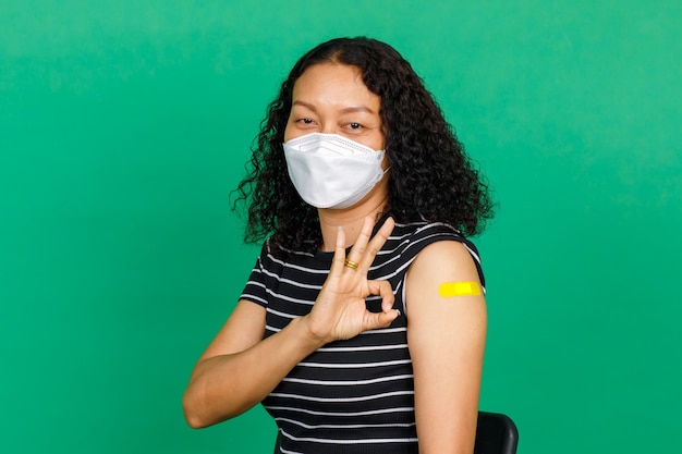 Asiatische Frau mittleren Alters mit Maske und Daumen hoch, ihr Arm mit Verbandspflaster zeigt, dass sie auf grünem Hintergrund gegen das Covid-19-Virus geimpft wurde. Konzept für die Covid-19-Impfung.