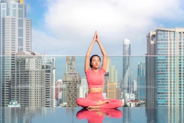 Asiatische Frau posiert beim Yoga auf dem Dach eines Hotels mit Blick auf die Stadt Bangkok und den Swimmingpool