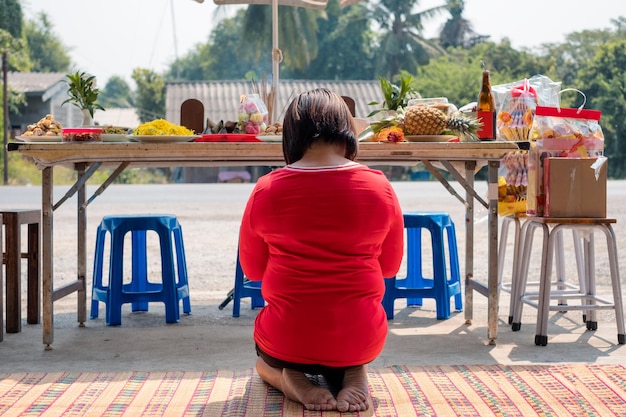 Asiatische Frau rotes Hemd sitzt und betet zum Vorfahren mit Mahlzeitdessert und Getränk auf dem Tisch in Neujahrschinesisch