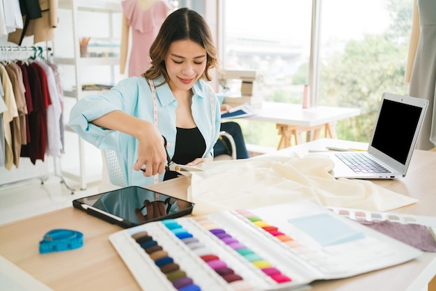 Asiatische Frau Schneiderin oder Modedesignerin schneidet Stoff mit einer Schere, um ein Muster von Kleidung zu machen Modedesignerin Schneiderin oder Näherin in der Werkstatt, die für neue Kollektionskleidung arbeitet