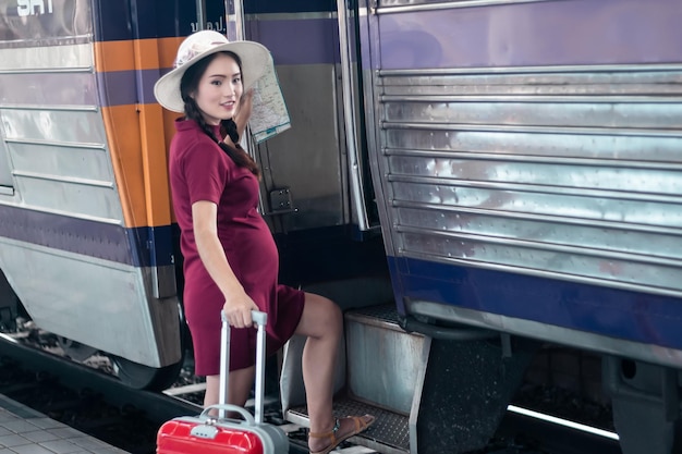 Asiatische Frau schwanger im roten Kleid mit einer Karte mit rotem Koffer am Bahnhof traveltraveler mit Rucksack im Sommer Urlaubskonzept Thailand