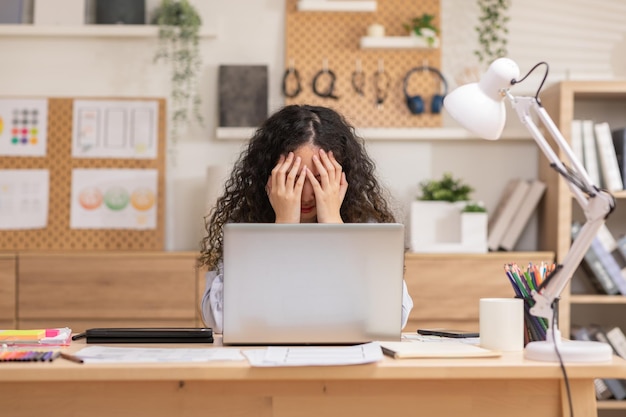 Asiatische Frau sitzt an einem Schreibtisch vor einem Laptop, ist gestresst, ihr Gesicht und ihre Kopfschmerzen zeigen, dass sie mit der Arbeit von zu Hause aus gestresst ist.
