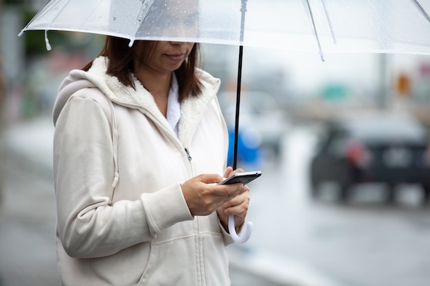 Asiatische Frau verwendet auf Smartphone, prüft Social-Media-Netzwerk und hält Regenschirm, während Taxi auf der Stadtstraße am regnerischen Tag wartet.