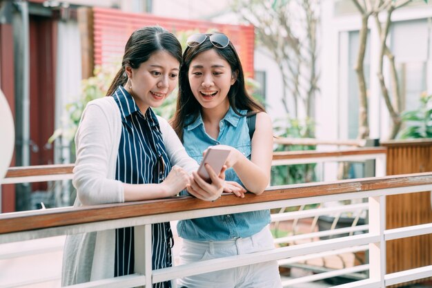 asiatische frau zeigt auf den bildschirm des smartphones ihrer freundin, während sie im sommer spaß haben, bilder an einem geländer in einem gemütlichen außenbereich zu betrachten.