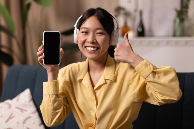 Asiatische Frau zeigt Smartphone-Bildschirm mit Kopfhörern und gestikuliert wie drinnen