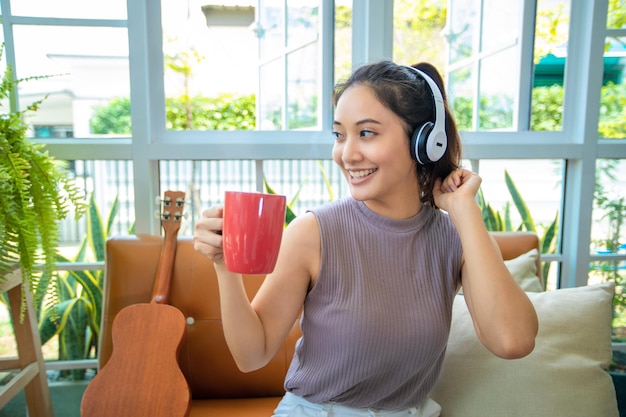 Asiatische Frauen, die Kopfhörer tragen und Laptop und digitales Tablet verwenden, um an einem entspannten Tag Musik zu hören und zu singen und zu Hause Kaffee zu trinken