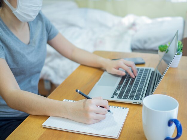 Asiatische Frauen, die Masken tragen, arbeiten zu Hause mit dem Laptop und schreiben in ein Notizbuch, um die Verbreitung des Coronavirus zu verringern