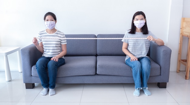 Asiatische Frauen, die Masken tragen, sitzen im Abstand voneinander auf dem Sofa und halten ein Maßband zur sozialen Distanzierung, um die Ausbreitung des Coronavirus zu verringern