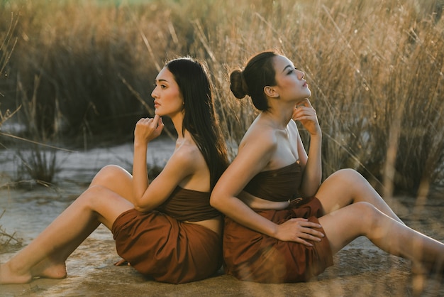 Asiatische Frauen Drama-Stil Kleid thailändische Tracht tragen. Porträt der schönen Freundin, die am Strand mit Grasfeldsonnenuntergangsnatur sitzt