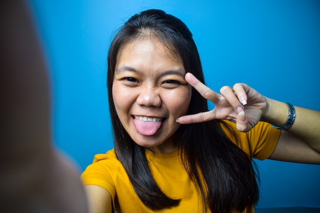 Asiatische Frauen mit blauem, isoliertem Hintergrund lächeln, nehmen Selfie und machen Frieden oder Siegeszeichen