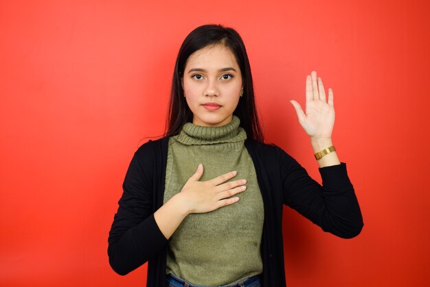 Asiatische Frauen mit schwarzem Pullover, die mit der Hand auf der Brust und der offenen Handfläche isoliert auf Rot schwören