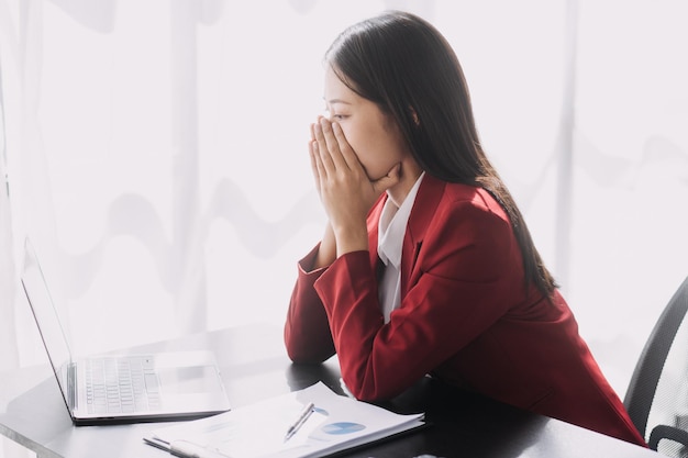 Asiatische Frauen sind gestresst, während sie am Laptop arbeiten Müde asiatische Geschäftsfrau mit Kopfschmerzen im Büro, die sich am Arbeitsplatz krank fühlt