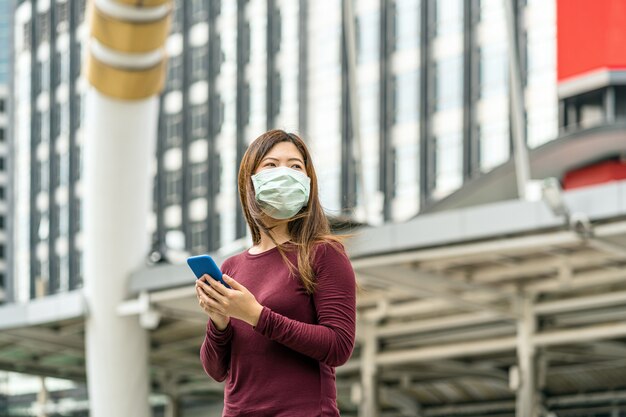 Asiatische Frauenhand mit intelligentem Mobiltelefon mit medizinischer Sicherheitsgesichtsmaske