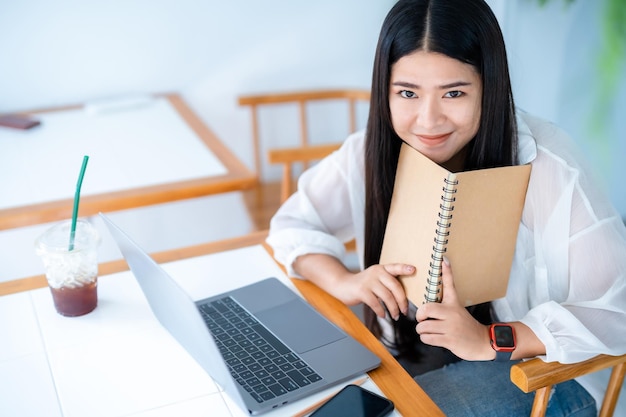 Asiatische, freiberuflich tätige Geschäftsfrauen, die mit einem Laptop arbeiten, um im Internet zu surfen, zu chatten und zu bloggen, und ein Smartphone-Notebook in einem öffentlichen Café