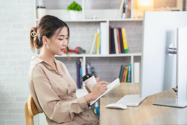 Asiatische freiberufliche Frau lächelt, hält eine Tasse heißen Kaffee und arbeitet zu Hause an einem Laptop-Computer auf einem Holztisch. Unternehmerin, die zu Hause für ihr Unternehmen arbeitet