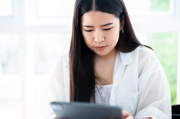 Asiatische freiberufliche Geschäftsfrau mit Smartwatch im Gespräch, die Gelegenheitsarbeit mit Tablett zum Surfen im Internet beim Chatten und Bloggen und Smartphone-Notebook im Café verwendet