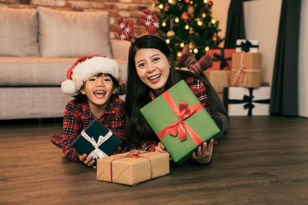 asiatische fröhliche familie mit geschenkboxen, die auf dem boden liegen und in die kamera schauen. junge mutter und schöne tochter zeigen die weihnachtsgeschenke. Frohe Weihnachten und ein frohes neues Jahr kommen!