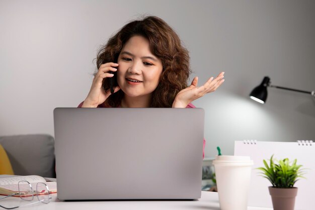 Asiatische Geschäftsfrau arbeitet zu Hause mit Laptop und spricht auf dem Smartphone und lächelt. Frau, die am Computer arbeitet und Handy benutzt, während sie zu Hause sitzt