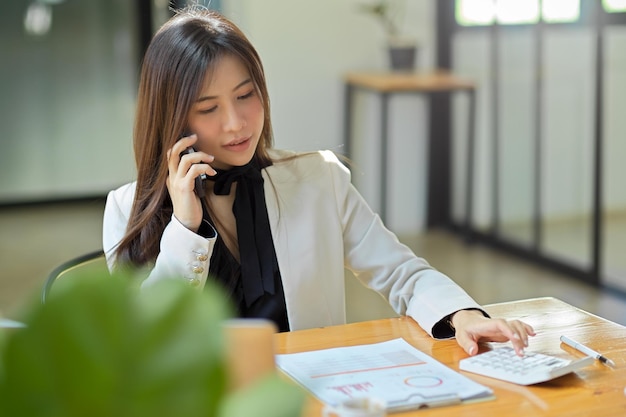 Asiatische Geschäftsfrau, die ein Smartphone hält und spricht, während sie einen Taschenrechner auf dem Schreibtisch benutzt