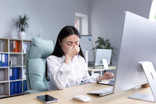 Asiatische Geschäftsfrau, die krank im Büro arbeitet und starke Kopfschmerzen hat, die Hände auf dem Kopf halten