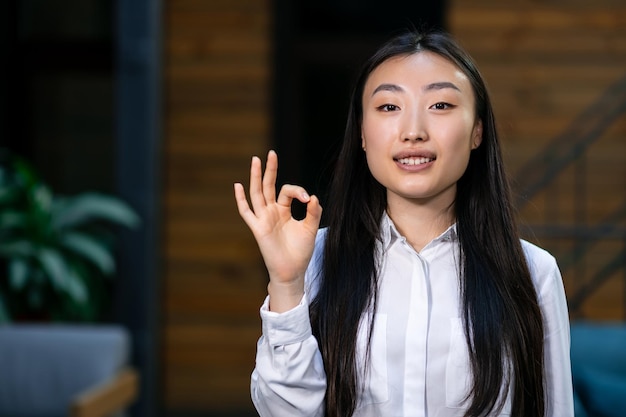 Asiatische Geschäftsfrau, die lächelt und ein OK-Schild zeigt, das auf dem Flur im Büro steht