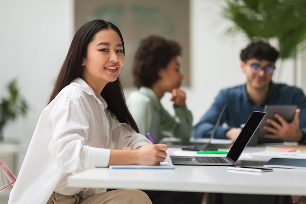 Asiatische Geschäftsfrau in der Nähe eines Laptops sitzt mit Kollegen drinnen am Schreibtisch