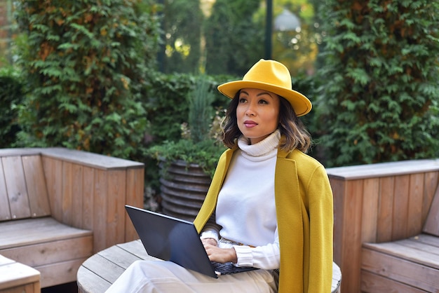 Asiatische Geschäftsfrau in einem gelben Mantel sitzt im Park und benutzt einen Laptop im Herbst