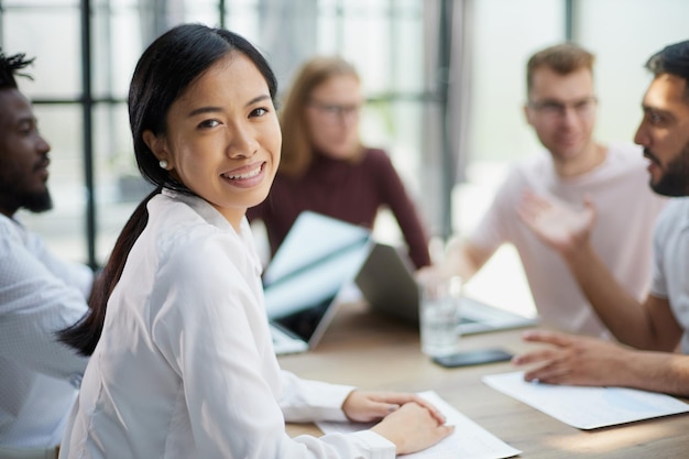 Asiatische Geschäftsfrau mit ihrer Mitarbeitergruppe im Hintergrund im modernen, hellen Büro im Innenbereich