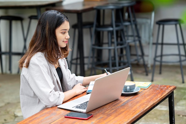 Asiatische Geschäftsfrau schreibt in ein Notizbuch mit einem Mobiltelefon-Laptop und einer Tasse Kaffee auf dem Tisch