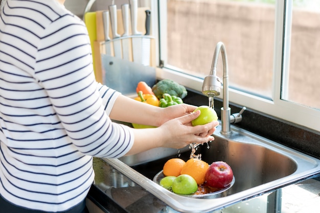 Asiatische gesunde Frau, die einen Apfel und andere Früchte über Küchenspüle wäscht und ein Obst / Gemüse mit Wasser reinigt, um das Risiko einer Kontamination zu beseitigen COVID-19.