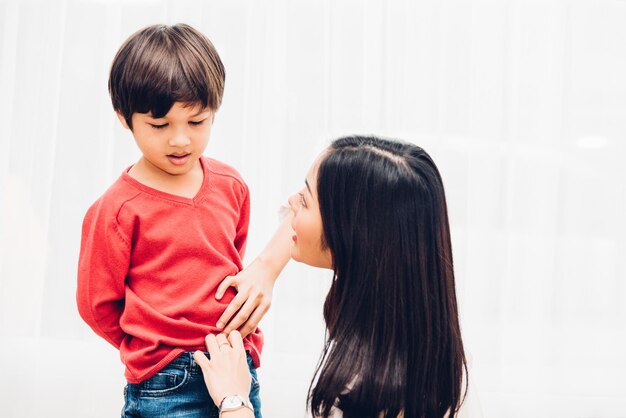 Asiatische, glückliche, schöne Mutter, die hilft, Kleidung für ihren Jungen im Haus zu bekommen, bevor sie zur Schule gehen, Zurück zum Schulkonzept