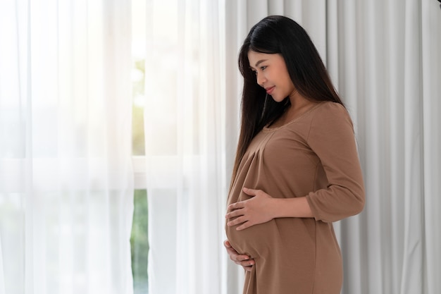 Asiatische glückliche schwangere Frau steht neben Fenstern und berührt ihren Bauch. Schwangerschaft, Mutterschaft, Menschen und Erwartungskonzept