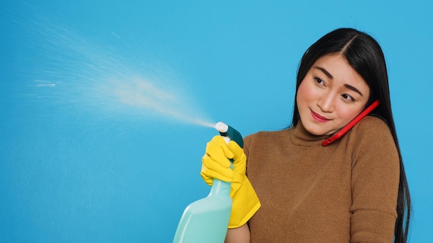 Asiatische Haushälterin, die chemisches Reinigungsmittel und Staubtuch verwendet, um das Haus zu reinigen, während sie mit entfernten Freunden am Handy telefoniert und entfernte Gespräche führt. Housekeeping- und Reinigungskonzept.