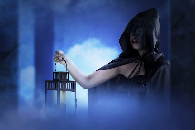 Asiatische Hexenfrau mit einem schwarzen Mantel, der eine Laterne hält, die in einem verlassenen Gebäude steht. Halloween-Konzept