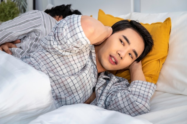 Asiatische homosexuelle Paare in den Pyjamas, die auf dem Schlafzimmer schnarchen und schlecht schlafen. Er, der Ohren mit den Händen blockiert